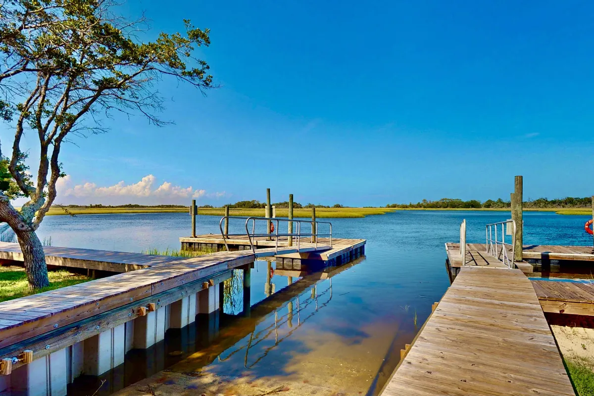 A scenic view of wooden docks stretching out into calm waters, surrounded by trees and open sky. The area is tranquil, perfect for leisurely strolls, enjoying sunset picnics, or relaxing by the water.