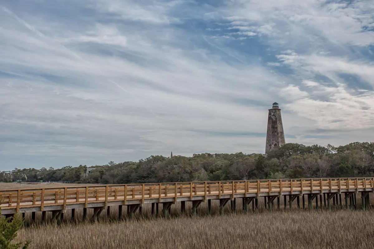 A picturesque view of a lighthouse stands at the edge of lush marshland, accessible via a wooden boardwalk. The area is surrounded by natural greenery and open sky, highlighting the island’s maritime heritage and ecological diversity.