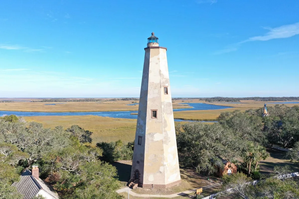 A striking view of a lighthouse surrounded by expansive marshland, a winding river, and open sky. The image is suffused with the golden light of sunset, with opportunities for birdwatching, photography, and soaking up the rich wildlife and scenery of Bald Head Island.