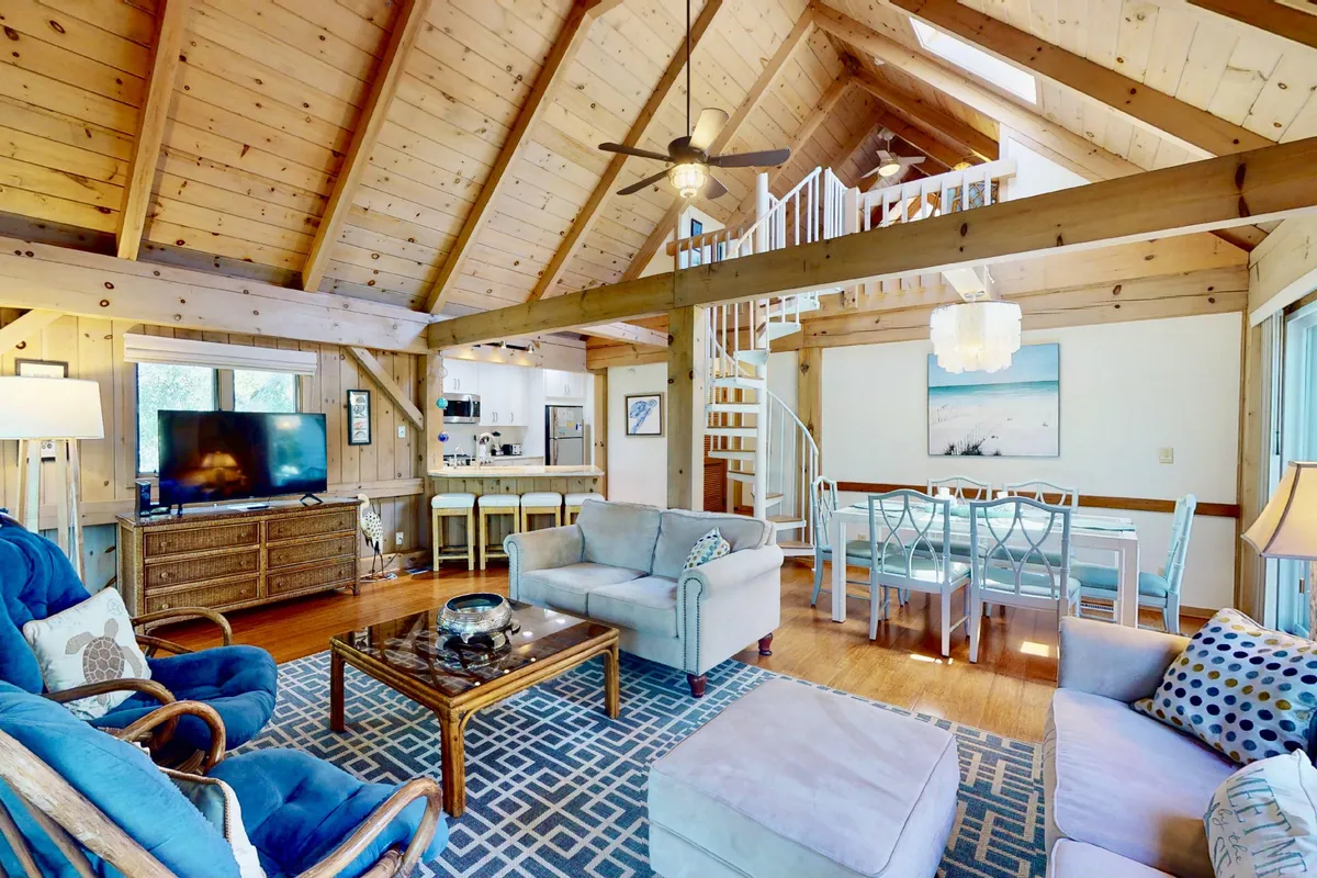 A bright, open-plan living and dining space with exposed wooden beams, large windows that frame the marsh view, a charming dining area, and a comfortable seating arrangement. Sunlight pours in through the windows, illuminating the decorative rug and inviting areas for relaxation and group activities.