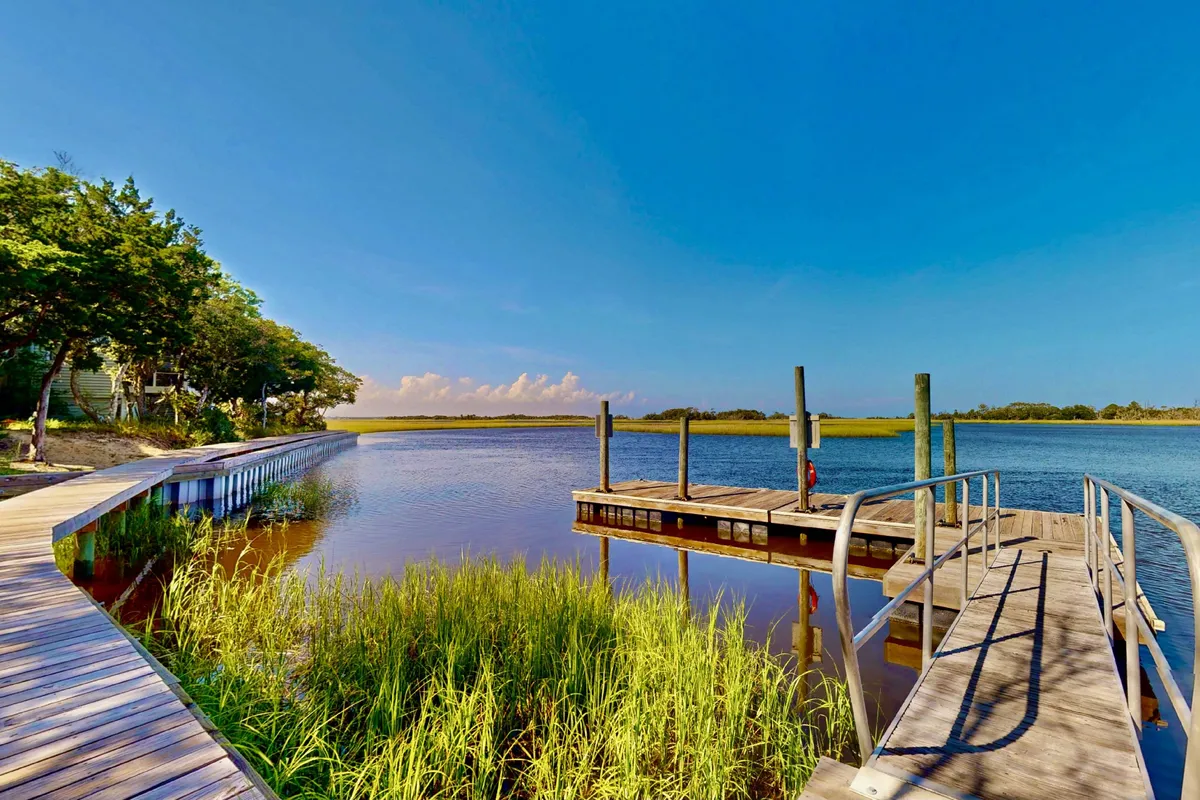 This image features a wooden dock extending over calm water, surrounded by lush grass and set against a clear blue sky. The dock offers easy access to aquatic activities and provides a peaceful spot to enjoy the tranquil marshlands.