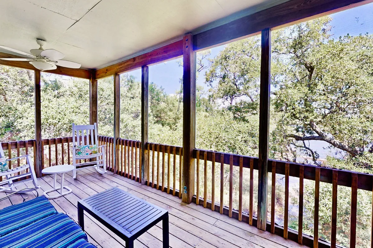This interior scene showcases a pair of classic wooden rocking chairs positioned in a screened-in porch, overlooking peaceful natural surroundings. A sturdy metal-framed table invites guests to gather, while expansive screened windows offer tranquil views perfect for birdwatching, enjoying coffee, or relaxing at sunset. The space feels bright, comfortable, and closely connected to nature.