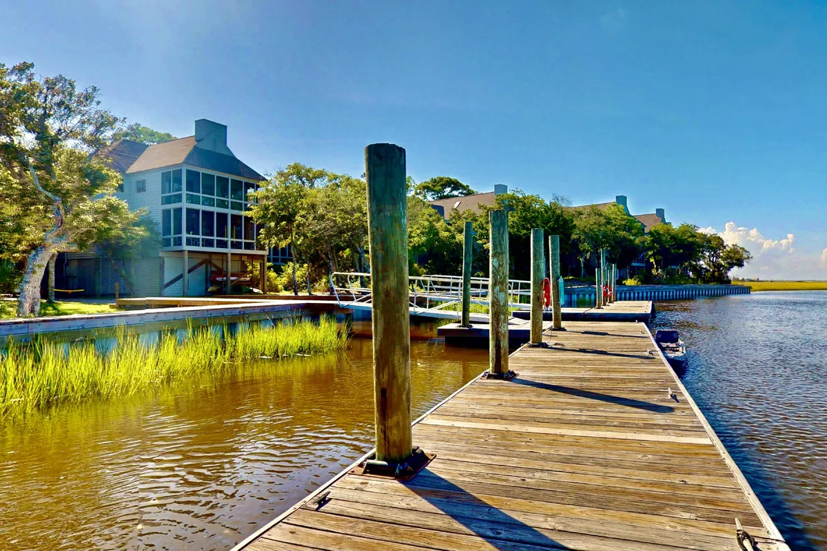 A wooden dock extends over calm water, bordered by lush marsh greenery. Buildings are visible in the distance, and the setting exudes tranquility, making it ideal for water-based activities like kayaking, fishing, and observing wildlife. The dock is the perfect launching point for marsh adventures directly from the property.