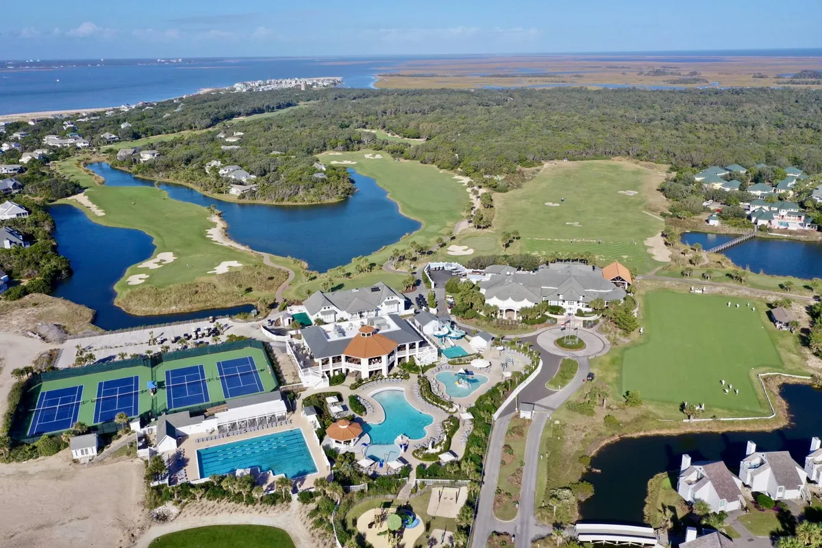 An aerial view encompassing a golf course, multiple swimming pools, tennis courts, residential buildings, and water features. The setting evokes a sense of resort-style leisure and a wide range of upscale amenities.