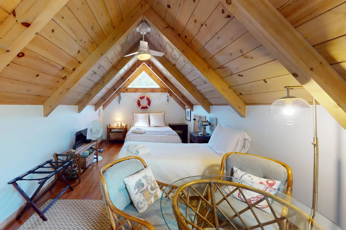 A charming loft space with an A-frame ceiling, a bed adorned with decorative pillows, and a small dining table with chairs nearby. The room glows with natural light from the window, creating a warm, multi-functional retreat ideal for kids or extra guests.