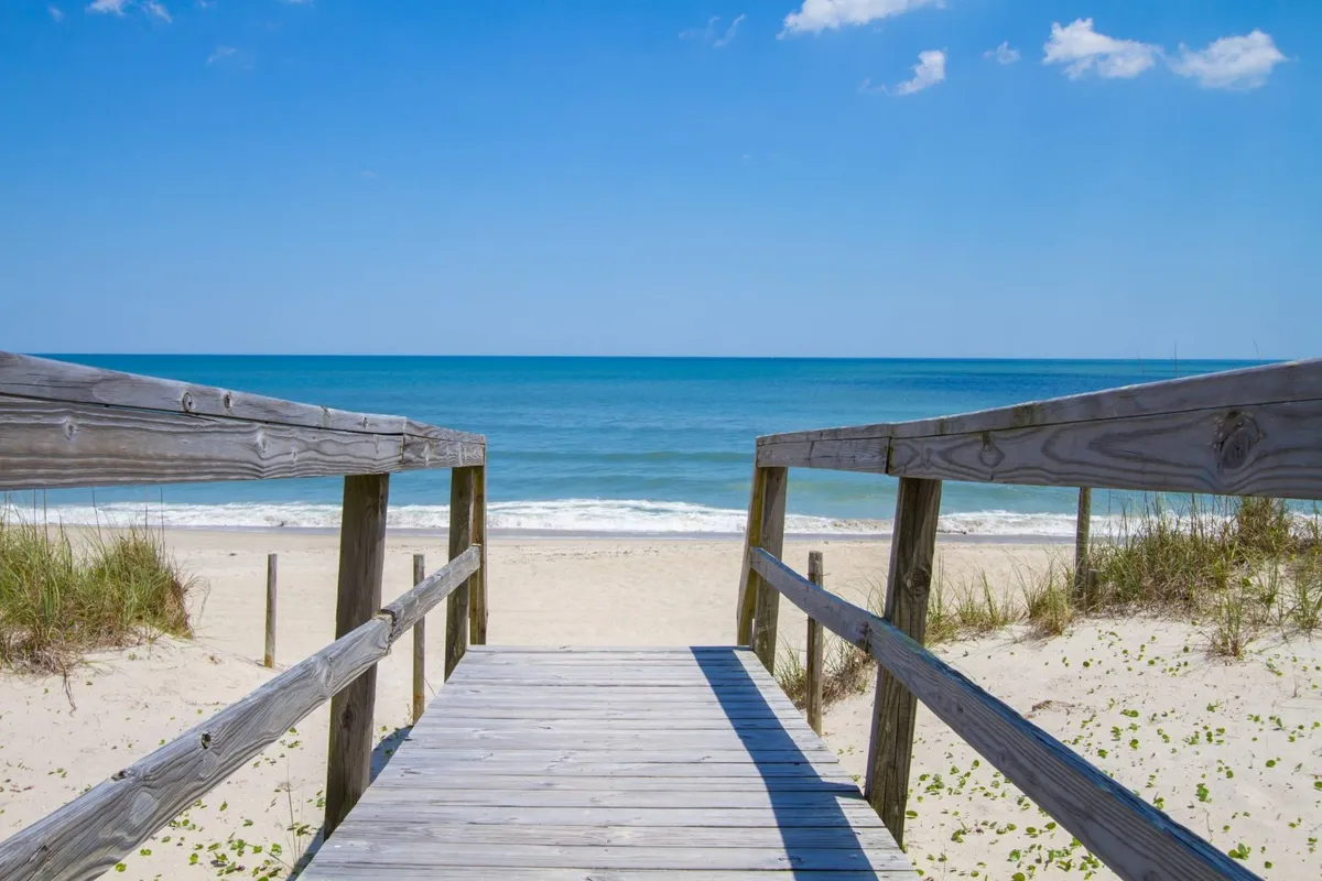 A wide wooden boardwalk leads directly to the sandy beach, bordered by dune grass and overlooking the sparkling ocean. The scene is open and peaceful, inviting visitors toward the shoreline for activities like beachcombing, sunbathing, or simply enjoying the view.