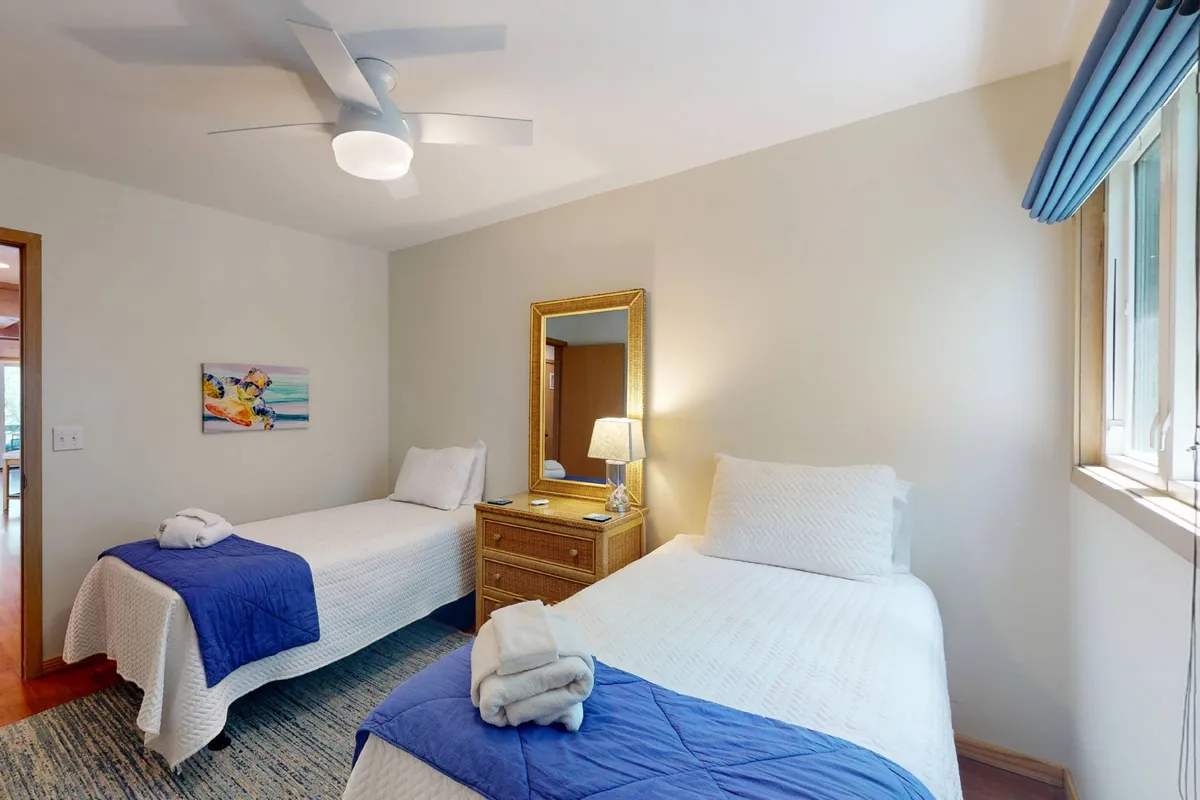 A cozy bedroom with two twin beds adorned in blue accents, a golden-framed mirror, a nightstand with a lamp between the beds, and sunlight filtering in, creating a bright and inviting setting. The arrangement is ideal for children or friends traveling together.