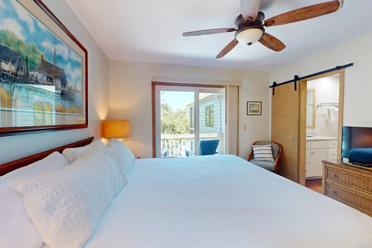 A spacious bedroom featuring a large, plush bed (likely the main suite’s king), tasteful artwork on the wall, a window inviting ample natural light, a ceiling fan, and an ensuite bathroom visible through an open door. The overall atmosphere is serene and comfortable.