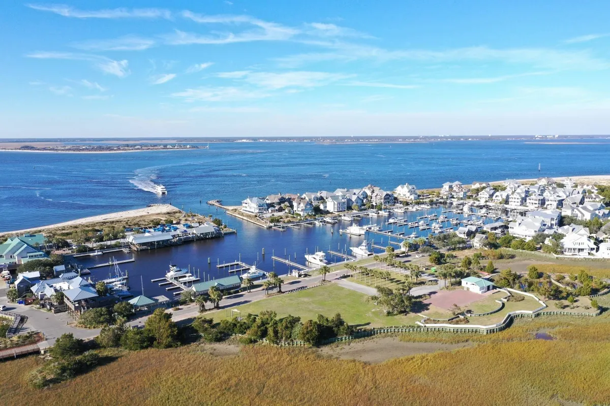 A panoramic view of a marina with waterfront houses, boats, lush greenery, and a broad expanse of water. The lively yet serene setting reflects a sense of community and resort living.